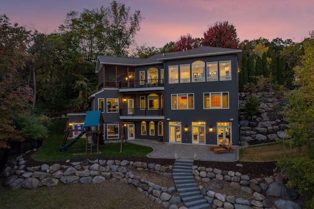 back house at dusk featuring a patio area, a playground, and a balcony