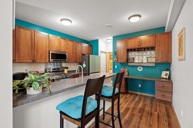 kitchen with a kitchen bar, brown cabinets, stainless steel appliances, a peninsula, and dark wood-style flooring