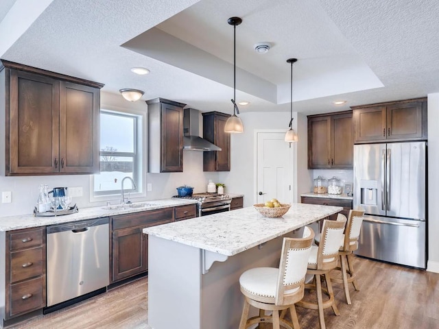 kitchen with a center island, wall chimney exhaust hood, sink, hanging light fixtures, and appliances with stainless steel finishes