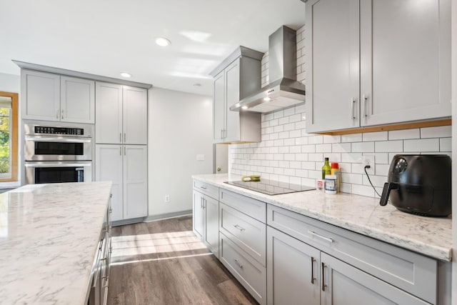 kitchen featuring stainless steel double oven, black electric cooktop, wall chimney exhaust hood, dark wood-type flooring, and light stone countertops