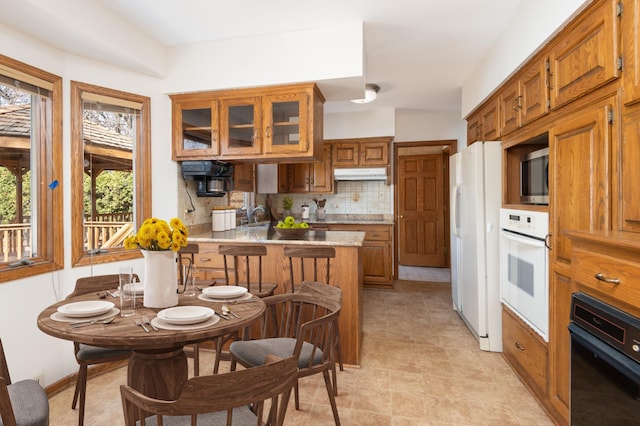 kitchen with brown cabinets, under cabinet range hood, tasteful backsplash, white appliances, and a peninsula