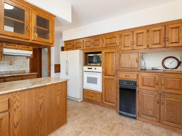 kitchen with tasteful backsplash, under cabinet range hood, light stone counters, brown cabinets, and white appliances