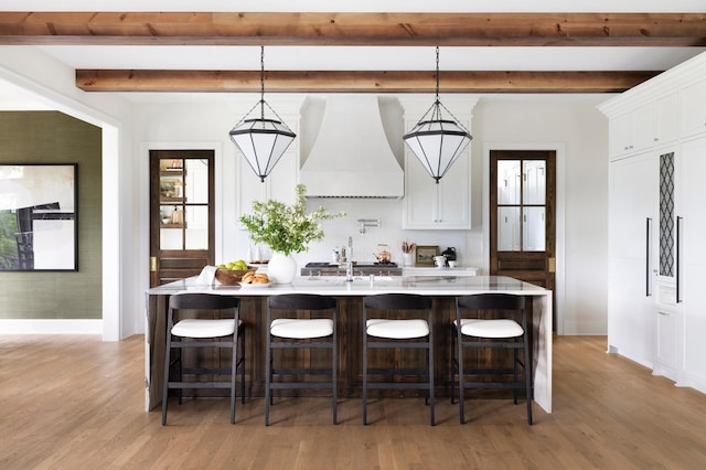 kitchen with white cabinetry, hanging light fixtures, premium range hood, a kitchen bar, and a kitchen island with sink