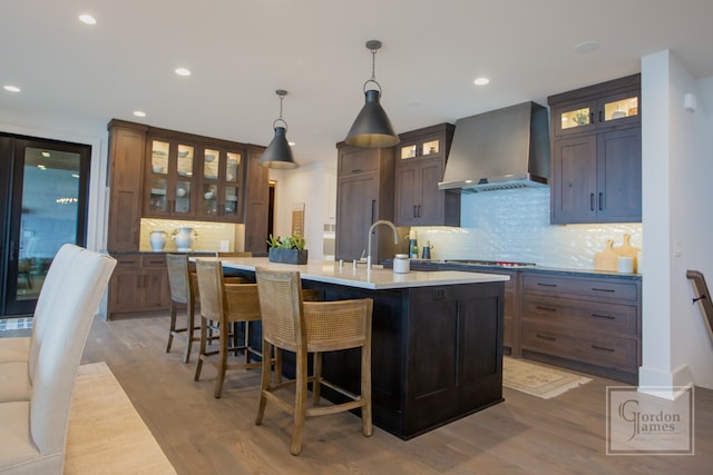 kitchen featuring pendant lighting, wall chimney exhaust hood, a kitchen island with sink, and wood-type flooring