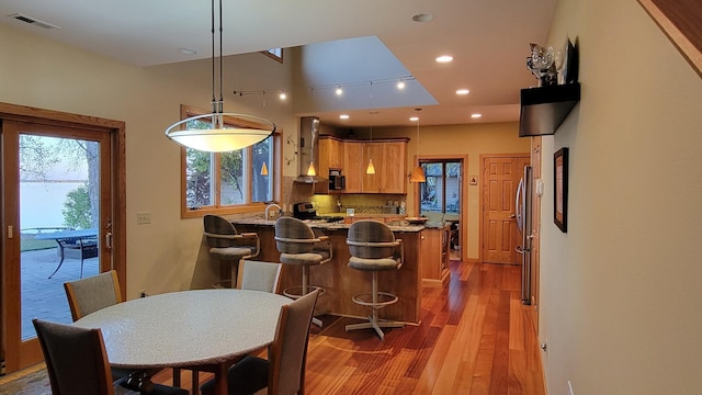 kitchen featuring hardwood / wood-style floors, backsplash, appliances with stainless steel finishes, a kitchen breakfast bar, and wall chimney exhaust hood