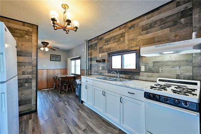 kitchen with white appliances, wooden walls, dark wood-style floors, a sink, and ceiling fan with notable chandelier