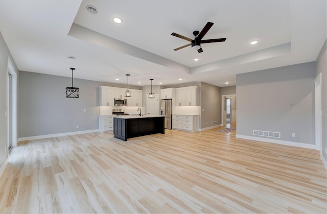 kitchen with pendant lighting, stainless steel appliances, an island with sink, white cabinets, and a raised ceiling