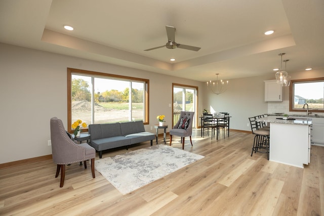 living room featuring ceiling fan with notable chandelier, light wood-type flooring, a tray ceiling, and sink