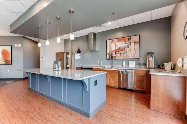 kitchen with light stone countertops, hanging light fixtures, stainless steel appliances, wall chimney exhaust hood, and light hardwood / wood-style flooring