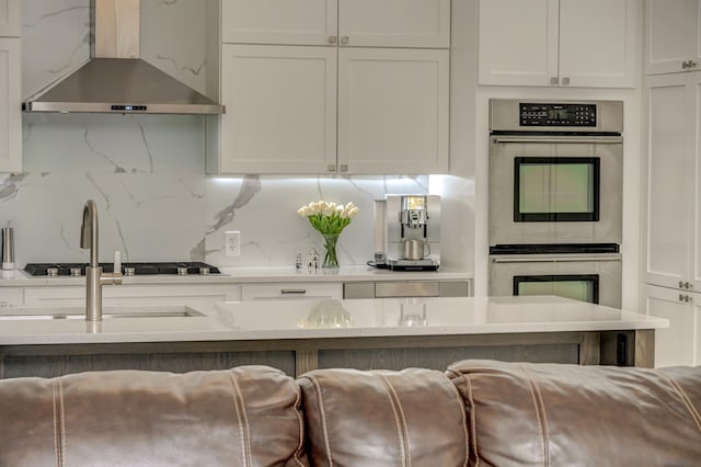 kitchen featuring wall chimney exhaust hood, stainless steel double oven, white cabinetry, and backsplash