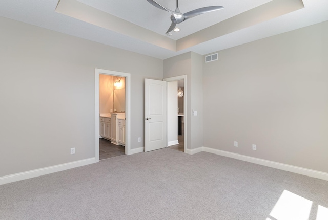 unfurnished bedroom with a raised ceiling, ensuite bathroom, ceiling fan, and light carpet