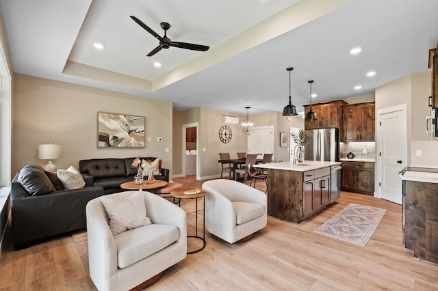 living room with ceiling fan with notable chandelier, a tray ceiling, sink, and light wood-type flooring