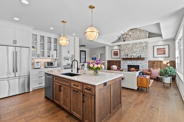 kitchen with a sink, light countertops, a stone fireplace, stainless steel appliances, and glass insert cabinets