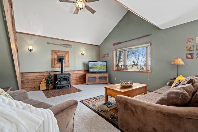 carpeted living room featuring ceiling fan, wooden walls, a wood stove, and high vaulted ceiling