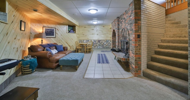 carpeted living room with stairway, wooden walls, a drop ceiling, and a brick fireplace