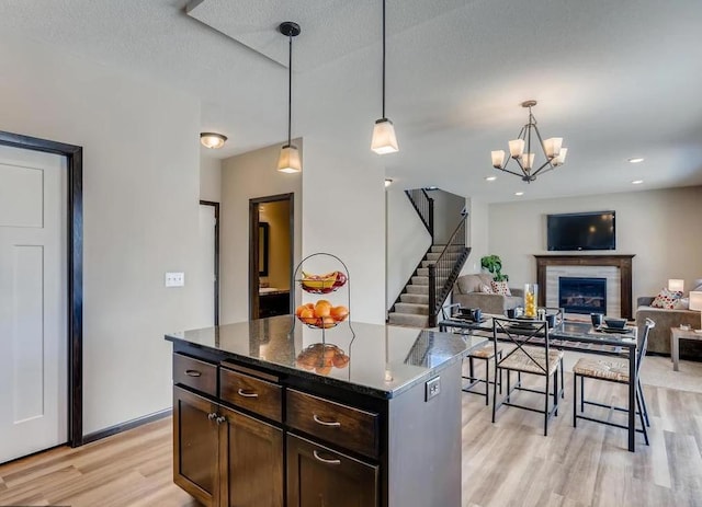 kitchen featuring dark brown cabinets, dark stone countertops, pendant lighting, a fireplace, and light hardwood / wood-style floors