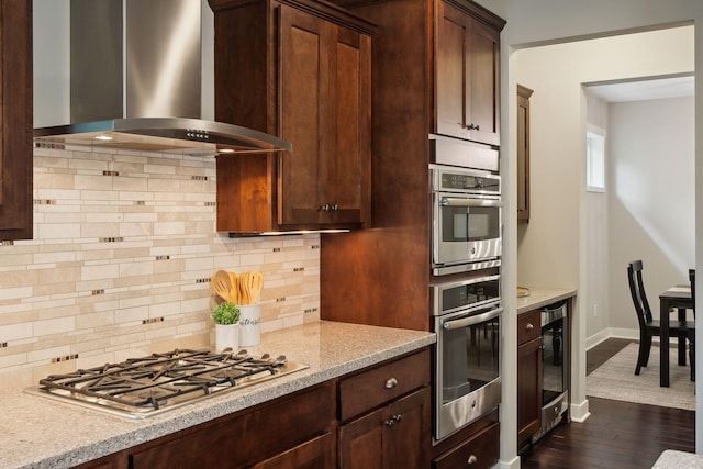 kitchen with wall chimney exhaust hood, dark wood-type flooring, stainless steel appliances, light stone counters, and tasteful backsplash