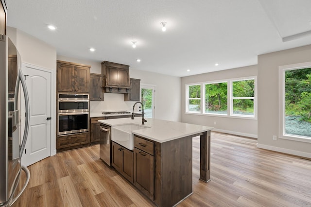 kitchen featuring appliances with stainless steel finishes, sink, dark brown cabinetry, light hardwood / wood-style flooring, and a center island with sink