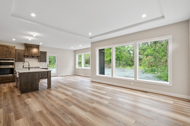 kitchen with a tray ceiling, a center island with sink, light wood-type flooring, and stainless steel double oven