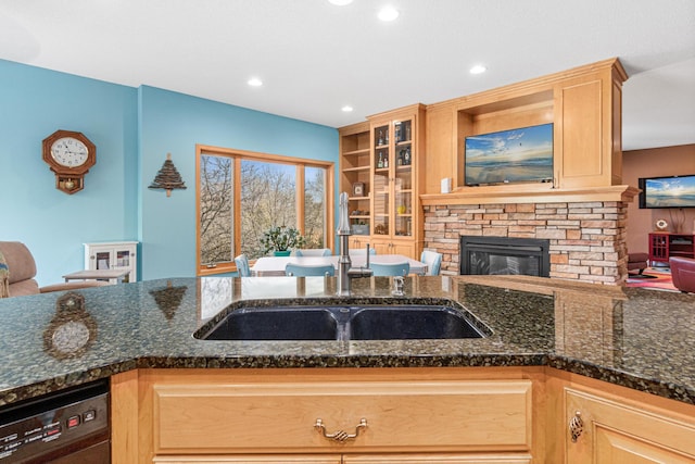 kitchen featuring sink, black dishwasher, a stone fireplace, dark stone counters, and light brown cabinetry