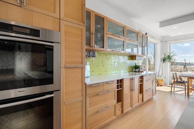 kitchen featuring stainless steel double oven, light countertops, light wood-type flooring, tasteful backsplash, and glass insert cabinets