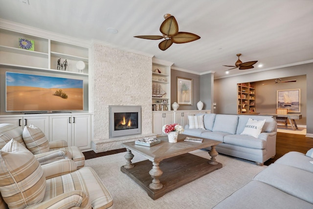 living room featuring light wood-type flooring, ornamental molding, built in shelves, ceiling fan, and a stone fireplace