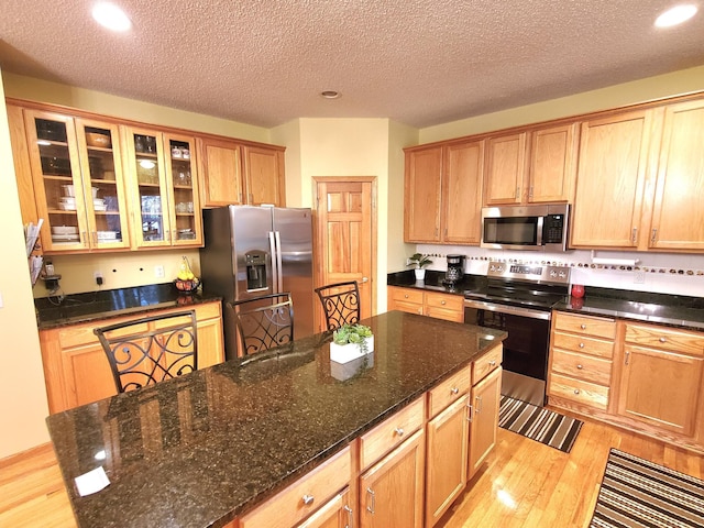 kitchen with a kitchen island, light wood-style floors, appliances with stainless steel finishes, dark stone counters, and glass insert cabinets