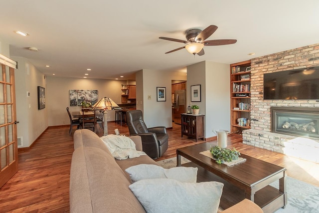 living room with a brick fireplace, built in shelves, hardwood / wood-style floors, and ceiling fan