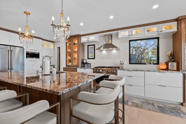 kitchen featuring a center island with sink, white cabinetry, hanging light fixtures, wall chimney exhaust hood, and high quality appliances