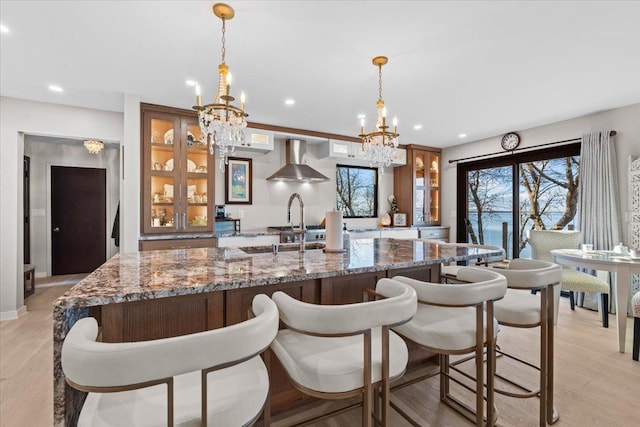 kitchen with wall chimney range hood, a chandelier, glass insert cabinets, a center island with sink, and recessed lighting
