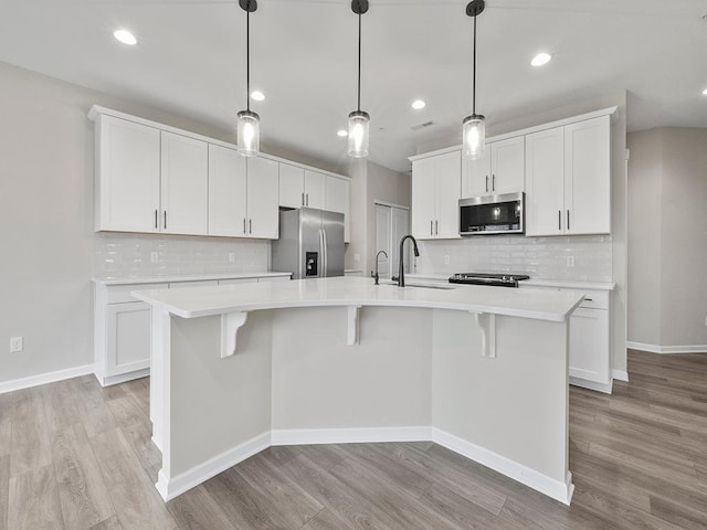 kitchen with light wood-type flooring, stainless steel appliances, pendant lighting, white cabinets, and an island with sink