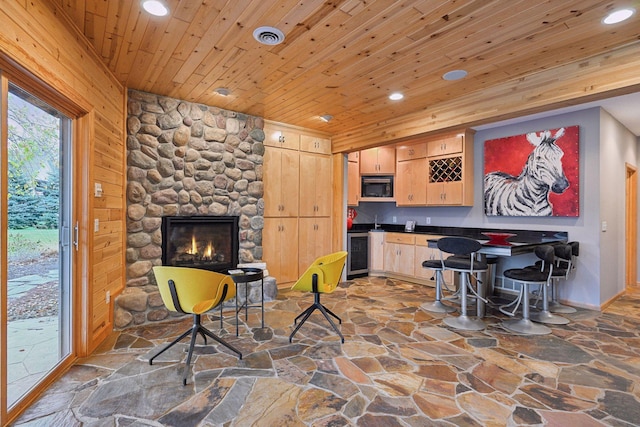 bar with a stone fireplace, black microwave, wooden ceiling, light brown cabinetry, and wine cooler