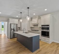 kitchen featuring wall chimney range hood, pendant lighting, a center island with sink, white cabinets, and appliances with stainless steel finishes