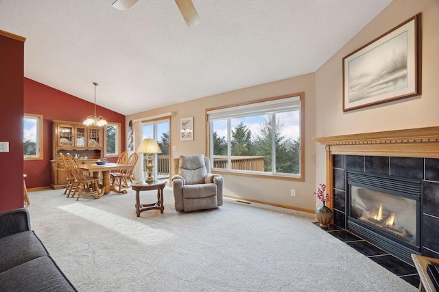 carpeted living area featuring ceiling fan with notable chandelier, visible vents, baseboards, vaulted ceiling, and a tiled fireplace