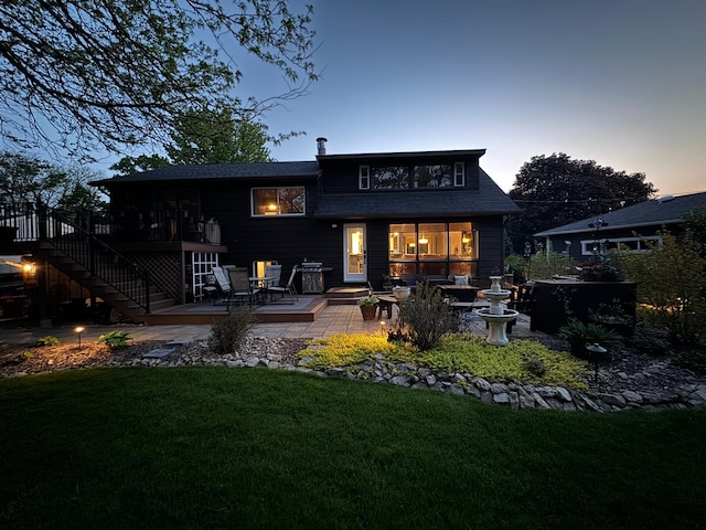 back house at dusk featuring a deck, a patio area, and a lawn