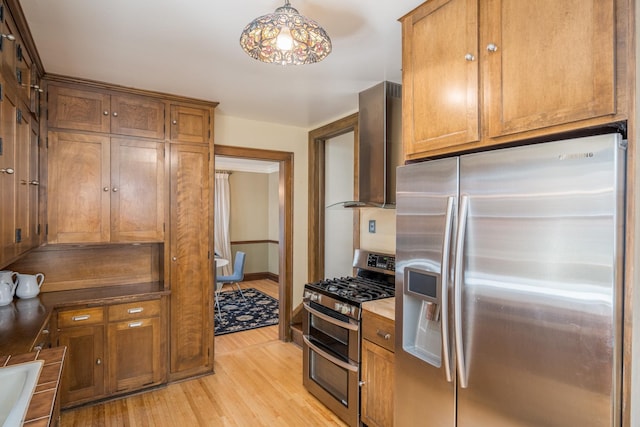 kitchen with hanging light fixtures, stainless steel appliances, wall chimney range hood, and light hardwood / wood-style flooring