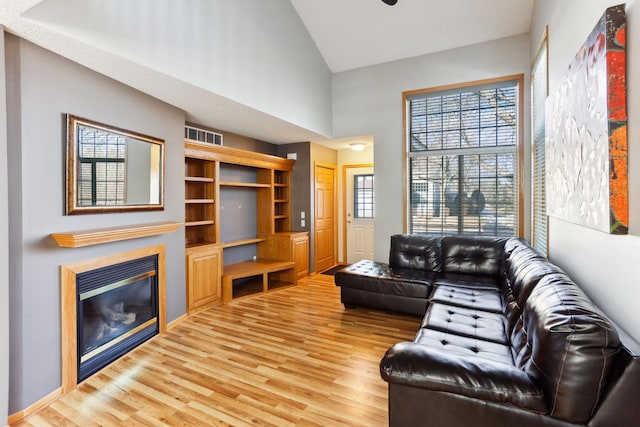 living room with a glass covered fireplace, light wood-style flooring, high vaulted ceiling, and visible vents