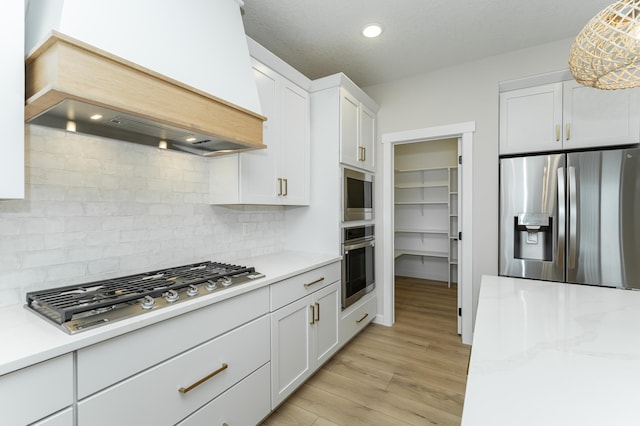 kitchen featuring premium range hood, white cabinets, appliances with stainless steel finishes, tasteful backsplash, and light hardwood / wood-style floors