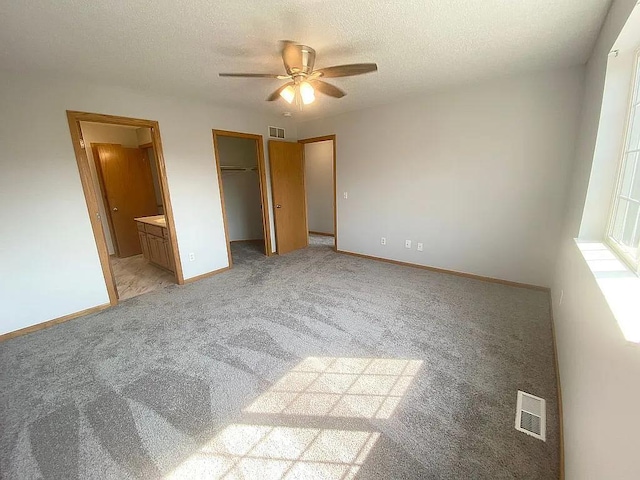 unfurnished bedroom featuring light carpet, ensuite bathroom, ceiling fan, a textured ceiling, and a walk in closet