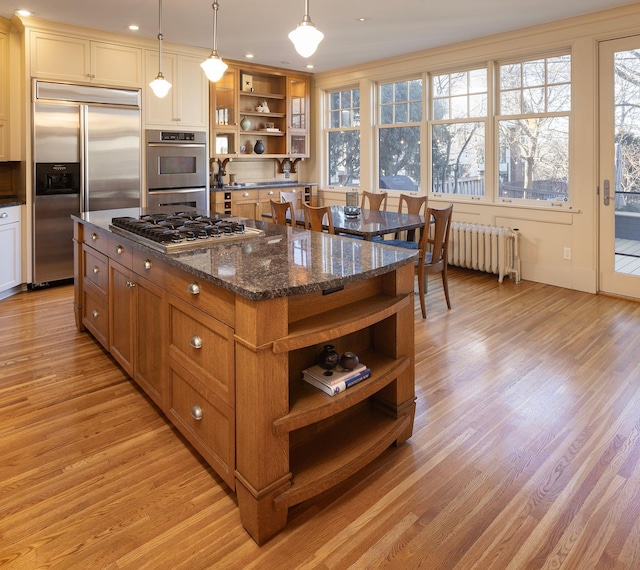 kitchen featuring radiator, stainless steel appliances, dark stone counters, decorative light fixtures, and light hardwood / wood-style floors