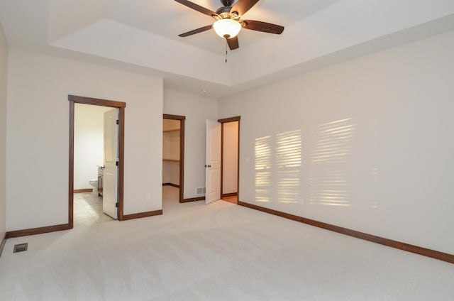 unfurnished bedroom featuring ensuite bathroom, ceiling fan, a raised ceiling, light carpet, and a walk in closet