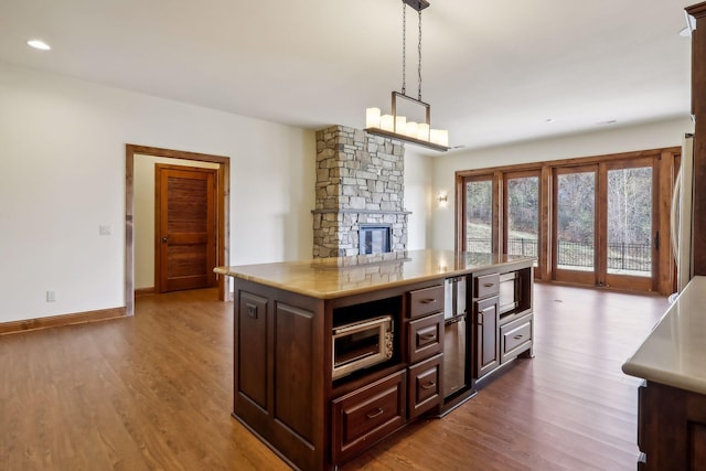 kitchen with a stone fireplace, built in microwave, hanging light fixtures, a center island, and dark brown cabinetry