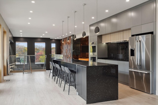 kitchen with sink, an island with sink, hanging light fixtures, stainless steel appliances, and light hardwood / wood-style flooring