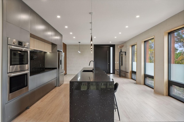 kitchen with sink, light wood-type flooring, an island with sink, hanging light fixtures, and stainless steel appliances