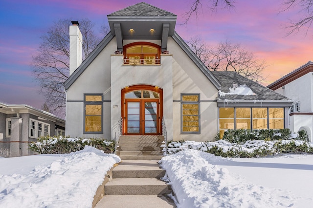 view of front facade featuring stucco siding, french doors, roof with shingles, a balcony, and a chimney