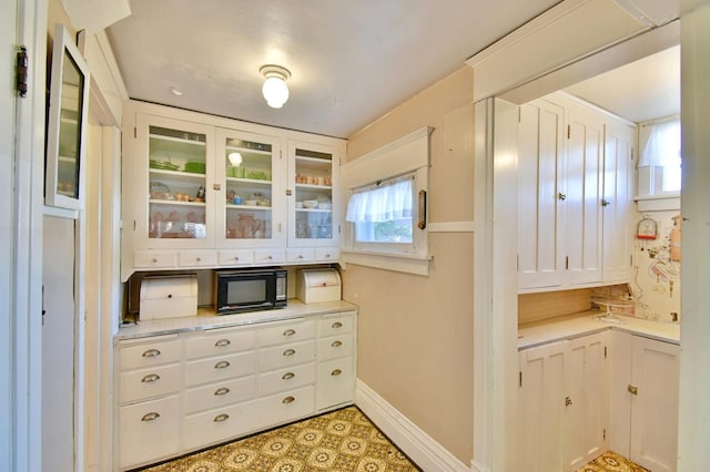 kitchen featuring black microwave, light countertops, baseboards, white cabinets, and glass insert cabinets