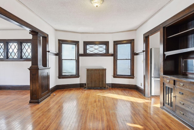 unfurnished living room with light wood-type flooring, ornate columns, radiator, ornamental molding, and a textured ceiling