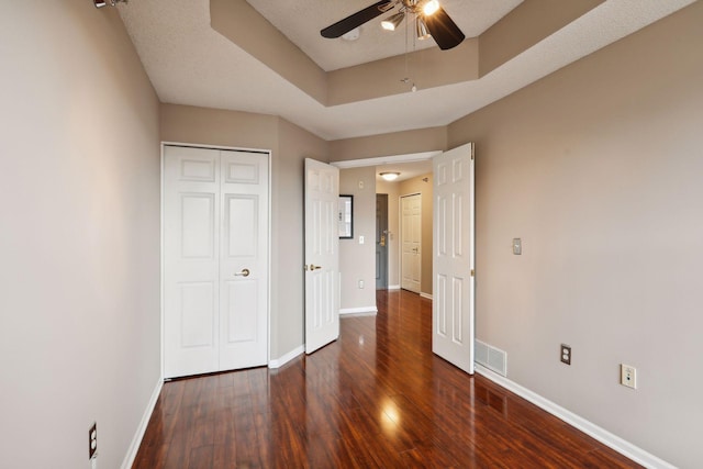 unfurnished bedroom featuring ceiling fan, a textured ceiling, a tray ceiling, dark hardwood / wood-style flooring, and a closet