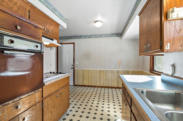 kitchen featuring a wainscoted wall, wallpapered walls, a sink, under cabinet range hood, and wall oven