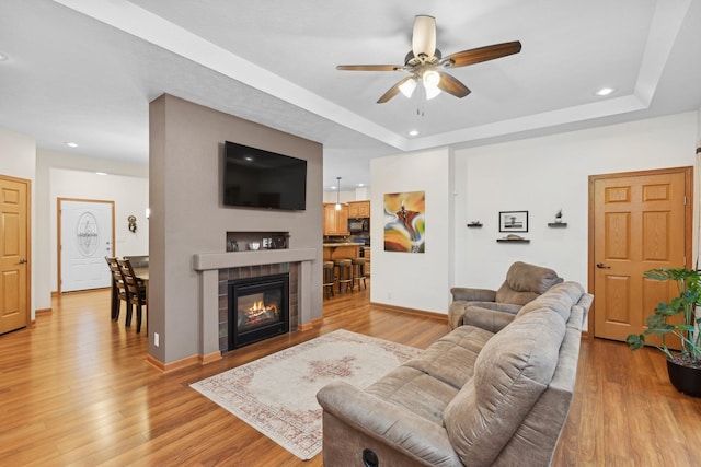 living room with light wood-type flooring, a raised ceiling, ceiling fan, and a tiled fireplace